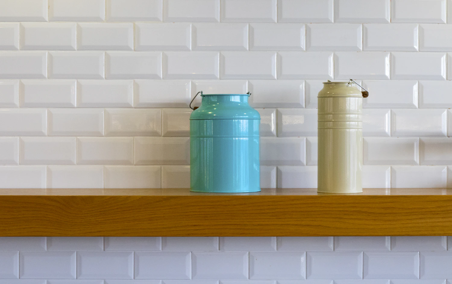 Two metal tins standing on a narrow wooden shelf against a white subway-tiled wall