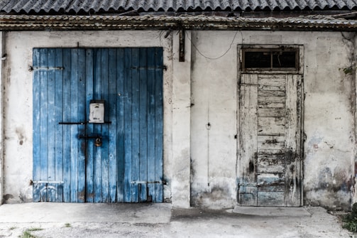 A weathered facade featuring two doors side by side, one painted blue with horizontal planks and a secure padlock, alongside a worn, unpainted wooden door with vertical slats. The surface around the doors is rough, adding to the rustic and aged appearance.