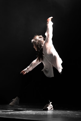 A performer is captured mid-movement in a dramatic spotlight, wearing a white shirt and dark pants. The stage is dimly lit, creating a stark contrast that emphasizes the dynamic pose.