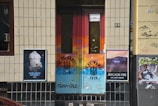 A colorful urban doorway with graffiti and posters on either side. The door features vibrant shades of orange and blue with a FC St. Pauli logo. To the left, there's a poster with a cow's head and German text, and to the right, a poster promoting an Arcade Fire album. The walls are tiled and bear some graffiti.