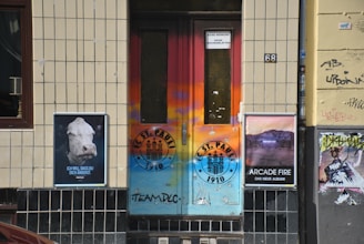 A colorful urban doorway with graffiti and posters on either side. The door features vibrant shades of orange and blue with a FC St. Pauli logo. To the left, there's a poster with a cow's head and German text, and to the right, a poster promoting an Arcade Fire album. The walls are tiled and bear some graffiti.