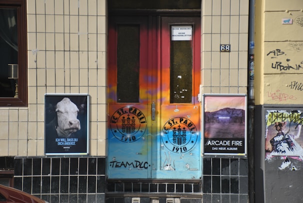 A colorful urban doorway with graffiti and posters on either side. The door features vibrant shades of orange and blue with a FC St. Pauli logo. To the left, there's a poster with a cow's head and German text, and to the right, a poster promoting an Arcade Fire album. The walls are tiled and bear some graffiti.