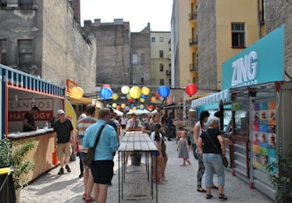 An outdoor market is situated between tall, worn buildings, featuring vibrant, colorful paper lanterns strung overhead. Stalls like 'Langos' and 'Zing' offer food and drinks to visitors. People of various ages are seen browsing, ordering snacks, and conversing in groups.