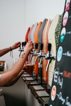 Several drink dispensers with various colored handles are lined up. A person is holding a plastic cup under one of the taps, filling it with a beverage. The foreground includes part of a blackboard menu with colorful writing and illustrations, indicating different drink options.