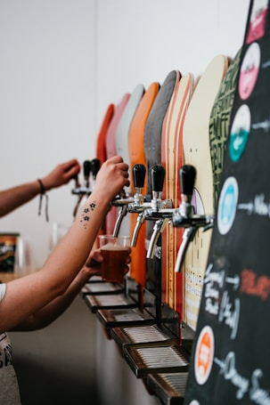 Several drink dispensers with various colored handles are lined up. A person is holding a plastic cup under one of the taps, filling it with a beverage. The foreground includes part of a blackboard menu with colorful writing and illustrations, indicating different drink options.