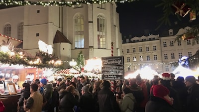 A bustling Christmas market with a crowd of people gathered around food stalls adorned with festive lights and decorations. The scene is set in front of a historic building, with a menu board displaying German food items. People are dressed warmly for the winter, some wearing festive hats, and there are strings of lights and decorations adding to the festive atmosphere.