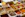 An assortment of colorful spices displayed in plastic containers, each with handwritten labels in a market setting. The spices range from vibrant reds and yellows to earthy browns and whites, accompanied by metal scoops for sampling.