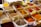 An assortment of colorful spices displayed in plastic containers, each with handwritten labels in a market setting. The spices range from vibrant reds and yellows to earthy browns and whites, accompanied by metal scoops for sampling.