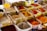 An assortment of colorful spices displayed in plastic containers, each with handwritten labels in a market setting. The spices range from vibrant reds and yellows to earthy browns and whites, accompanied by metal scoops for sampling.