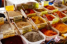 An assortment of colorful spices displayed in plastic containers, each with handwritten labels in a market setting. The spices range from vibrant reds and yellows to earthy browns and whites, accompanied by metal scoops for sampling.
