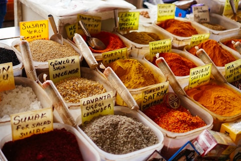 An assortment of colorful spices displayed in plastic containers, each with handwritten labels in a market setting. The spices range from vibrant reds and yellows to earthy browns and whites, accompanied by metal scoops for sampling.