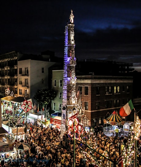 A vibrant street festival scene with a large crowd gathered around an ornate, illuminated tower structure adorned with intricate designs and topped by a statue. Colorful lights are strung along the street and there are various tents and stalls visible, including a small carousel. Buildings of mixed height surround the area, creating an enclosed festive atmosphere.