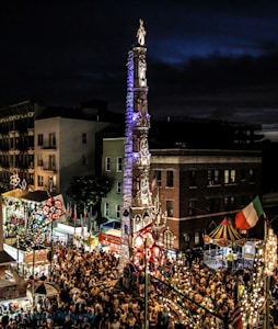 A vibrant street festival scene with a large crowd gathered around an ornate, illuminated tower structure adorned with intricate designs and topped by a statue. Colorful lights are strung along the street and there are various tents and stalls visible, including a small carousel. Buildings of mixed height surround the area, creating an enclosed festive atmosphere.