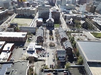 An aerial view of an urban area featuring a church with a prominent white spire at the center. Surrounding the church are residential buildings arranged symmetrically. To the side, there is a large parking lot and various commercial and residential structures. Streets with parked cars and some greenery are visible throughout the scene.