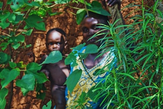 A woman and a child standing amidst dense green foliage. The woman is wearing a colorful dress and is partially hidden by leaves, while the child is naked and directly facing the camera. The background consists of a textured, reddish-brown mud wall.