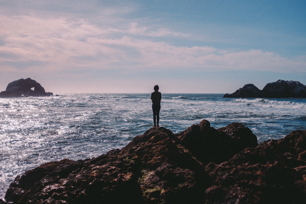 person standing on coastal rock facing ocean at daytime