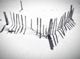 A series of wooden posts create a rustic fence in a sandy environment. The fence casts distinct shadows on the sand, with its posts appearing weathered and slightly uneven. Sparse footprints are visible in the surrounding sand, hinting at a recent presence.