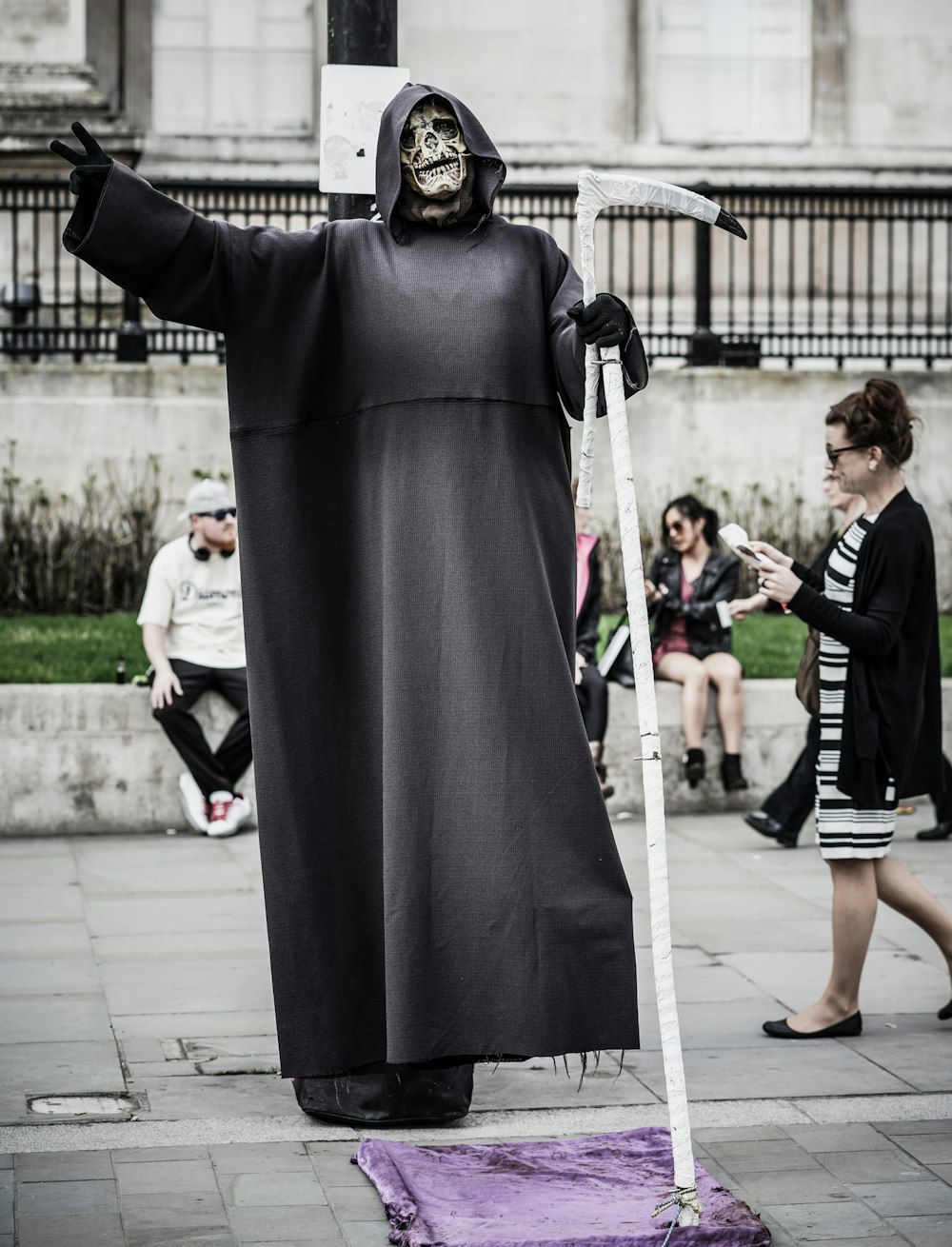 man in grim reaper costume with scythe near woman walking and people sitting on ledge