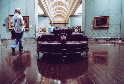 A spacious room in an art gallery features several paintings displayed on the walls. The central focus is a luxurious, dark leather circular bench on a shiny wooden floor. People are casually observing the artworks, creating a relaxed atmosphere.