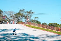 Several people are enjoying outdoor activities. One person is rollerblading downhill on a paved path, while others are engaged in a zip-line adventure above. A few onlookers stand nearby against a backdrop of lush green trees.