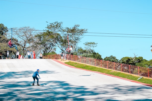 Several people are enjoying outdoor activities. One person is rollerblading downhill on a paved path, while others are engaged in a zip-line adventure above. A few onlookers stand nearby against a backdrop of lush green trees.