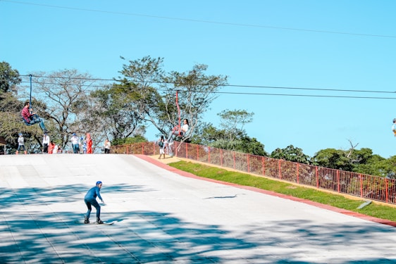 Several people are enjoying outdoor activities. One person is rollerblading downhill on a paved path, while others are engaged in a zip-line adventure above. A few onlookers stand nearby against a backdrop of lush green trees.