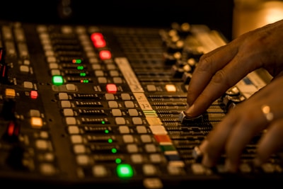 A close-up of a person's hands adjusting the sliders on an audio mixing console in a dimly lit environment. The console features numerous buttons, knobs, and sliders with various colored lights indicating different levels and functions.