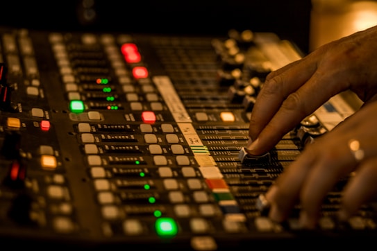 A close-up of a person's hands adjusting the sliders on an audio mixing console in a dimly lit environment. The console features numerous buttons, knobs, and sliders with various colored lights indicating different levels and functions.