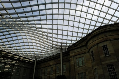 A large, curved glass roof structure covers a historic building, creating a modern architectural feature. The grid pattern of the glass panels allows natural light to filter through, casting a geometric shadow on the stone facade below.