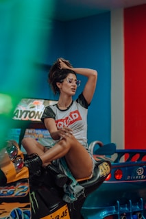 A person wearing sunglasses and a casual outfit sits confidently on an arcade racing game motorcycle. The setting appears to be a colorful game arcade with machines and vibrant lighting.