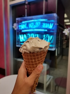 A hand holds a waffle cone filled with creamy ice cream, against a backdrop of a softly blurred neon sign in a window, suggesting a cozy indoor or cafe setting.
