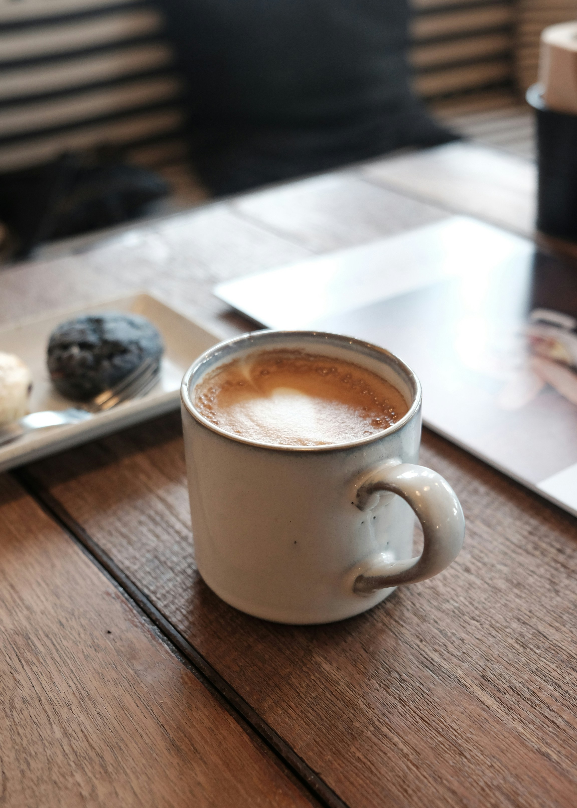 A ceramic mug filled with frothy coffee sits on a wooden tabletop. Nearby, a plate holds two snacks, possibly pastries or cookies. The setting appears to be a cozy cafe, with soft lighting and a relaxed ambiance. The background features striped seats and a partially visible magazine or book.