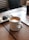 A ceramic mug filled with frothy coffee sits on a wooden tabletop. Nearby, a plate holds two snacks, possibly pastries or cookies. The setting appears to be a cozy cafe, with soft lighting and a relaxed ambiance. The background features striped seats and a partially visible magazine or book.