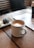 A ceramic mug filled with frothy coffee sits on a wooden tabletop. Nearby, a plate holds two snacks, possibly pastries or cookies. The setting appears to be a cozy cafe, with soft lighting and a relaxed ambiance. The background features striped seats and a partially visible magazine or book.