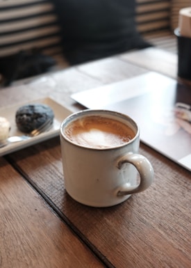 A ceramic mug filled with frothy coffee sits on a wooden tabletop. Nearby, a plate holds two snacks, possibly pastries or cookies. The setting appears to be a cozy cafe, with soft lighting and a relaxed ambiance. The background features striped seats and a partially visible magazine or book.