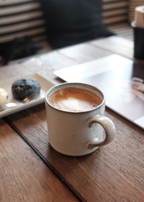 A ceramic mug filled with frothy coffee sits on a wooden tabletop. Nearby, a plate holds two snacks, possibly pastries or cookies. The setting appears to be a cozy cafe, with soft lighting and a relaxed ambiance. The background features striped seats and a partially visible magazine or book.