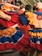 A group of people wearing vibrant, colorful traditional costumes with layered skirts in red, blue, and yellow hues. The attire is adorned with ruffles and patterns, and the scene appears lively and festive.