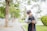 A young person with glasses is standing outdoors on a pathway, reading a book. They have headphones around their neck and hold a phone in one hand. The setting is a park-like environment with greenery and trees in the background.