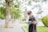 A young person with glasses is standing outdoors on a pathway, reading a book. They have headphones around their neck and hold a phone in one hand. The setting is a park-like environment with greenery and trees in the background.