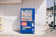 A bright blue vending machine stands against the exterior wall of a building. It is filled with a variety of drinks, including cans and bottles. On the right, a white truck is parked nearby. The background includes a closed garage door and light-colored wall.