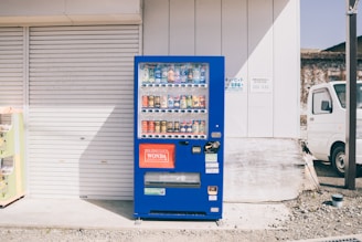 A bright blue vending machine stands against the exterior wall of a building. It is filled with a variety of drinks, including cans and bottles. On the right, a white truck is parked nearby. The background includes a closed garage door and light-colored wall.