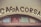 The image features a vintage-style sign with the word 'CASACORSA' in dark, bold letters. The sign is set above a textured archway with decorative stained glass panels in shades of blue, red, and green, creating a rustic and charming appearance.