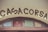 The image features a vintage-style sign with the word 'CASACORSA' in dark, bold letters. The sign is set above a textured archway with decorative stained glass panels in shades of blue, red, and green, creating a rustic and charming appearance.