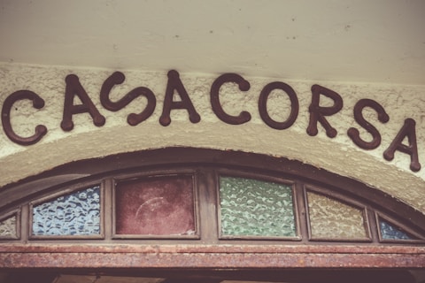 The image features a vintage-style sign with the word 'CASACORSA' in dark, bold letters. The sign is set above a textured archway with decorative stained glass panels in shades of blue, red, and green, creating a rustic and charming appearance.