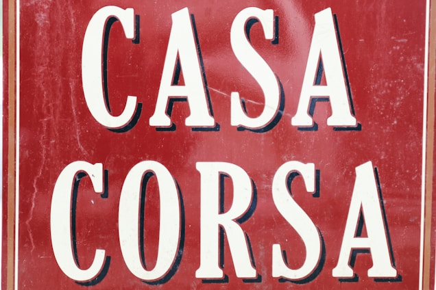 A vintage-style sign displays the words 'CASA CORSA' in bold, white letters with a black shadow effect. The background is a deep red, with visible signs of wear such as scratches and scuffs, adding a rustic and aged appearance. The overall layout is symmetrical, with the words stacked one above the other, framed with thin white and black borders.
