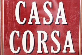 A vintage-style sign displays the words 'CASA CORSA' in bold, white letters with a black shadow effect. The background is a deep red, with visible signs of wear such as scratches and scuffs, adding a rustic and aged appearance. The overall layout is symmetrical, with the words stacked one above the other, framed with thin white and black borders.
