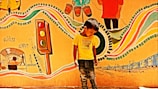 A young boy stands in front of a vibrant mural. The mural features colorful elements such as a traffic light, musical instruments, and abstract patterns. There are various depictions of urban life, including rickshaws and people walking, creating an energetic and lively scene.