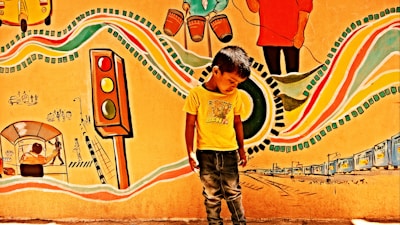 A young boy stands in front of a vibrant mural. The mural features colorful elements such as a traffic light, musical instruments, and abstract patterns. There are various depictions of urban life, including rickshaws and people walking, creating an energetic and lively scene.