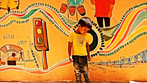 A young boy stands in front of a vibrant mural. The mural features colorful elements such as a traffic light, musical instruments, and abstract patterns. There are various depictions of urban life, including rickshaws and people walking, creating an energetic and lively scene.