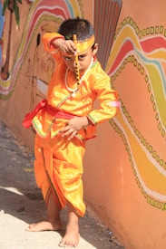 A young child is dressed in vibrant traditional attire standing in front of a colorful mural. The child is wearing an orange costume with a red sash and a headband, holding a flute in front of their face. The mural in the background features wave-like patterns in orange, yellow, and pink.